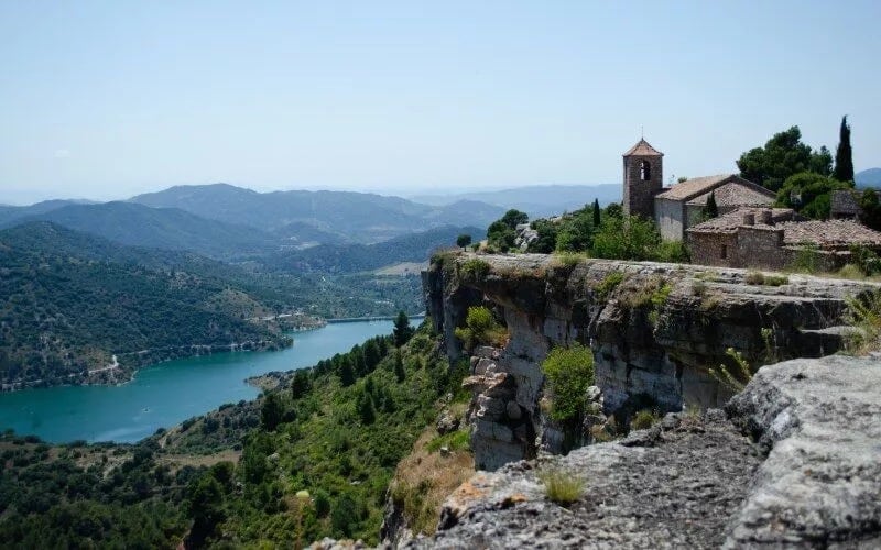 A village on the brink of a cliff, looking over a landscape with a river and mountains