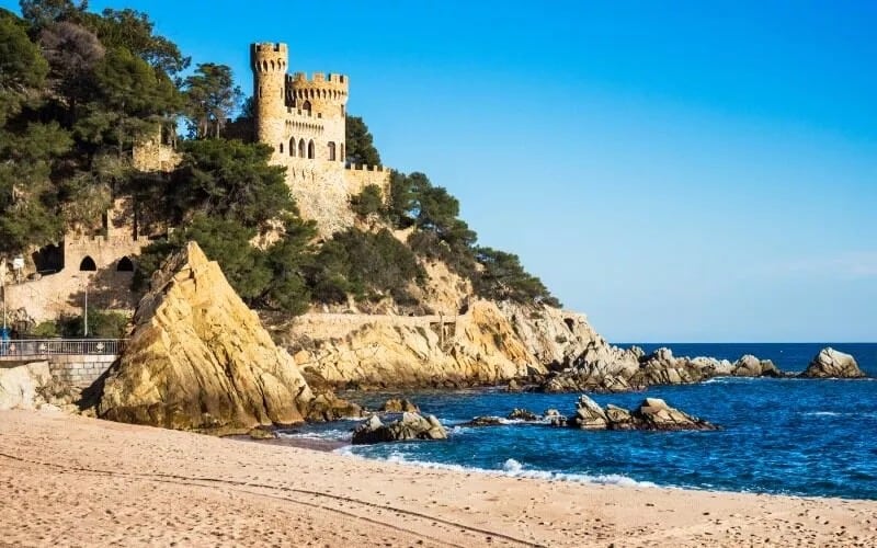 A castle on the coast surrounded by pine trees, and the sand of the beach in front
