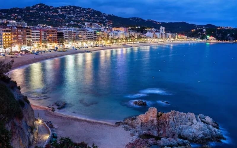 A beach and lit buildings reflecting on the water