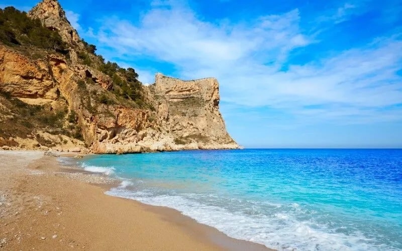 A beach with blue water and rocky cliffs on a side
