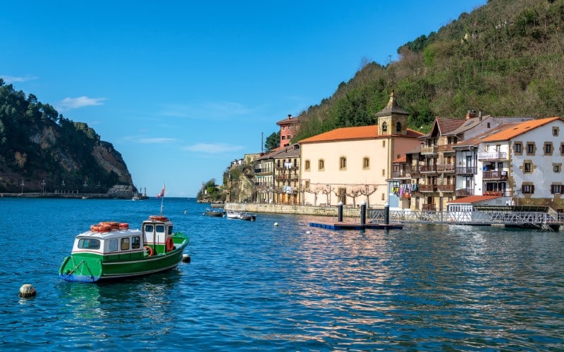 A green and white boat crossing a blue estuary towards a colourful village