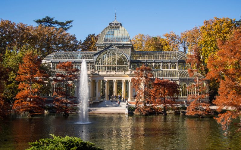 A glass palace in front of a pond surrounded by russet trees